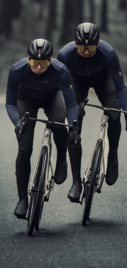 Two cyclists wearing black helmets and gear are riding on a road.