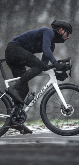A cyclist rides a white S-Works bike in snowy weather, wearing dark cycling gear.