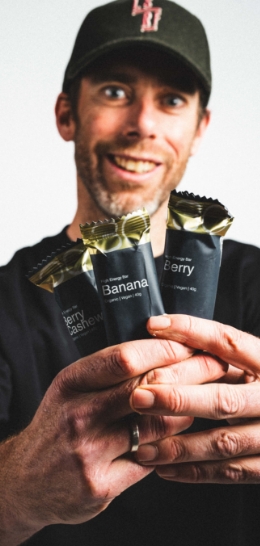 A man wearing a cap is holding three energy bars with visible labels: Berry Cashew, Banana, and Berry.