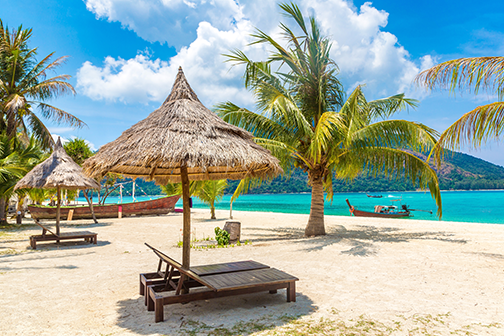Twee houten strandbedjes onder een rieten parasol op het strand met palmbomen en een bootje op de achtergrond op Aruba