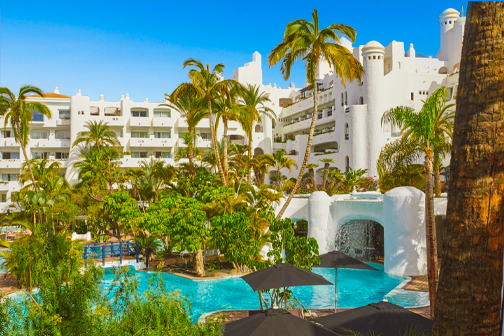 Zwembad en groene tuin met veel palmen en wit hotelgebouw