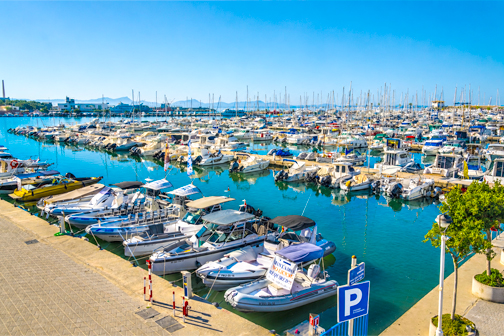 Haven van Port d'Alcúdia op Mallorca met zeilboten en jachten aan de steigers, helderblauw water en palmbomen langs de boulevard.