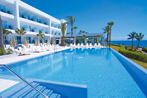 nfinity-zwembad van Riu Palace Meloneras Resort met ligbedden, palmbomen en uitzicht op zee onder een heldere blauwe lucht.