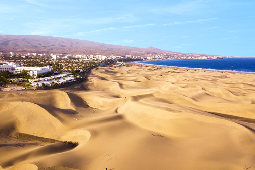 Glooiende zandduinen van Maspalomas op Gran Canaria met op de achtergrond een kustplaats, bergen en de zee onder een heldere lucht.