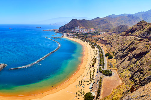 Uitzicht op een azuurblauwe baai op Gran Canaria met lang zandstrand, palmbomen, boulevard en bergen langs de kustlijn.