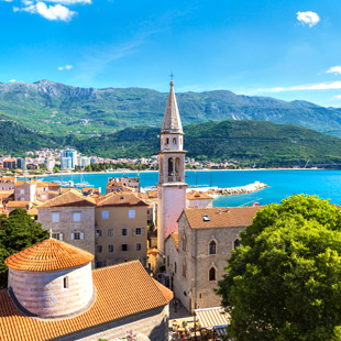 Centrum van Budva met oranje daken, kerktoren, blauwe zee en groene bergen op de achtergrond.